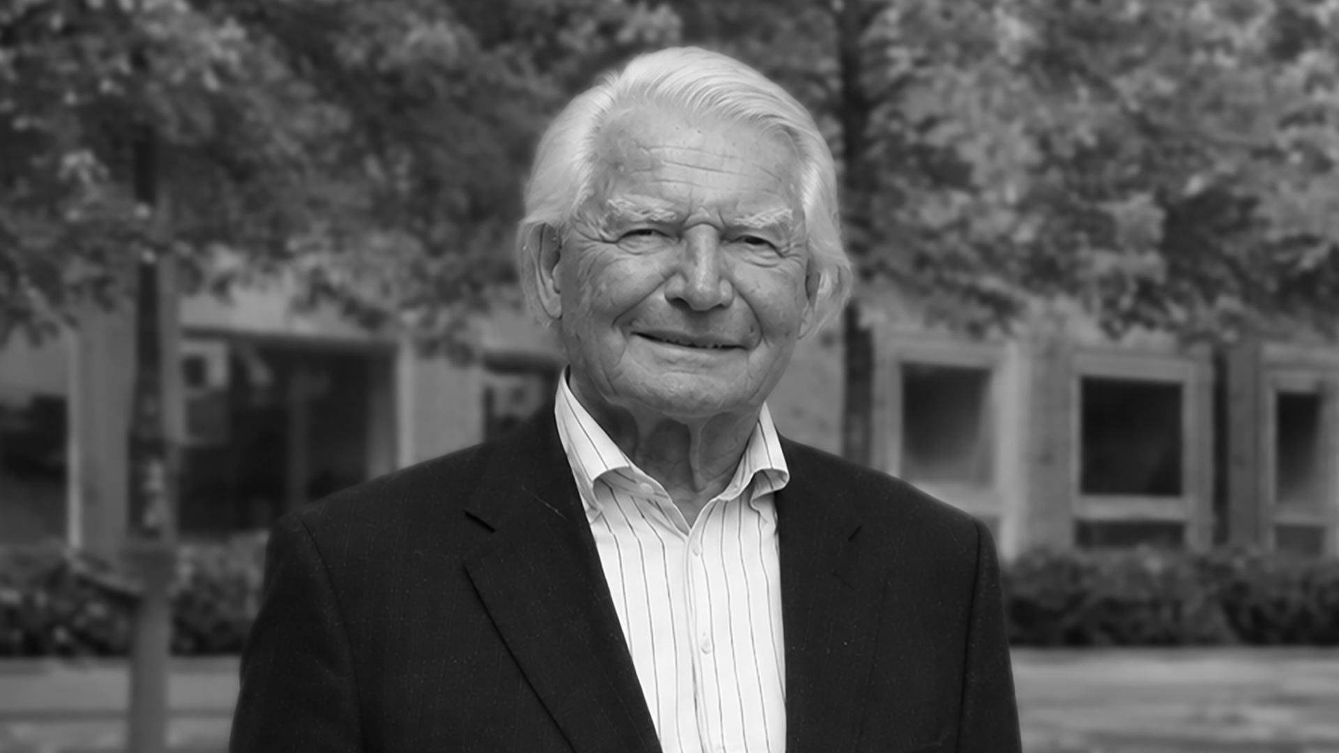 Elderly man in a suit smiles outdoors with trees and buildings in the background.